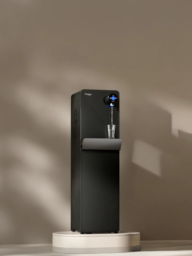 The Purity Prime, a black floor-standing dispenser pouring a glass of water, on a low plinth in a sunlit room.