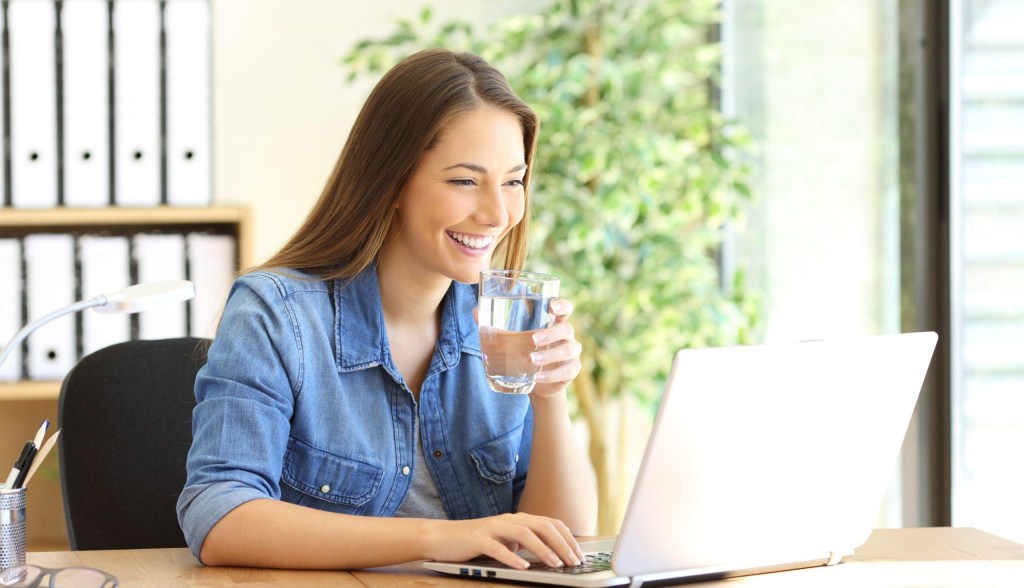 woman smiling while working on computer drinking water