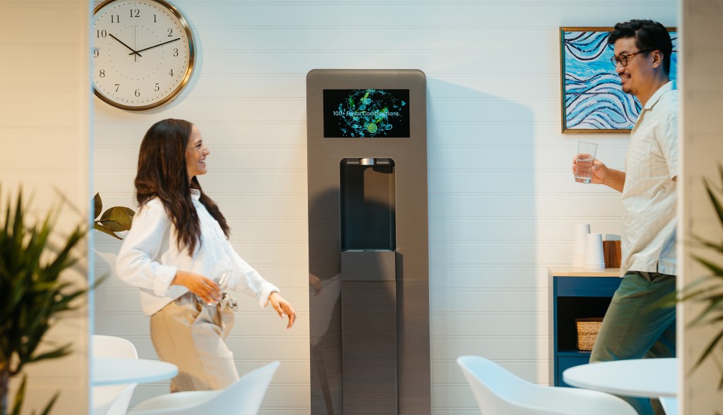 Man and woman with glass of water in hand walking toward Twist water dispenser smiling