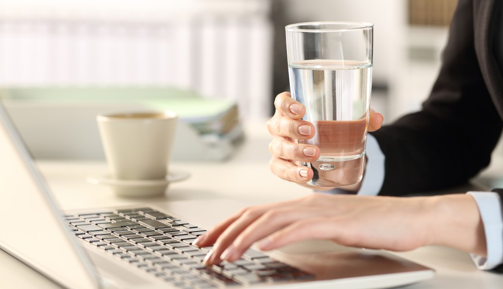 hand holding a glass of water in front of laptop with a mug in the background